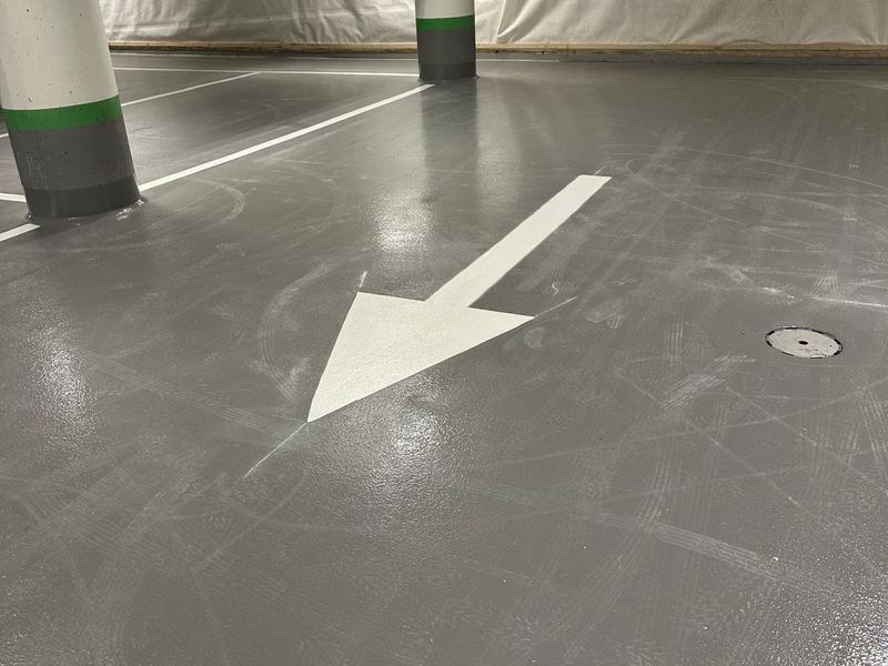 Vit riktningspil på ett grått garagegolv, med parkeringslinjer och pelare i bakgrunden.